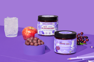 Two jars of 'Elderberry' supplement on a purple surface with an apple, berries, and a glass of water.